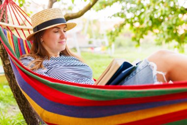 Yeşil bahçede rahat bir hamakta uzanmış kitap okuyan genç bir kadın..