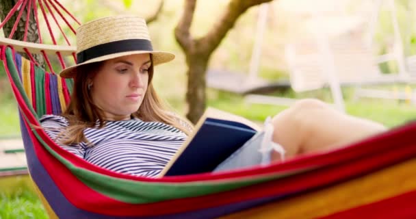 緑の庭で快適なハンモックに寝そべって本を読んでいる若い女性 高品質4K映像 — ストック動画
