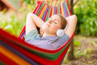 Kulaklıklı genç bir kadın dışarıda hamakta dinlenirken müzik dinliyor..