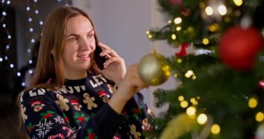 Genç bir kadın, Noel ağacını dekore ederken arkadaşlarıyla ya da akrabalarıyla neşeli bir oturma odasında cep telefonuyla konuşuyor. Noel 'i ve yeni yılı kutluyoruz. Yüksek kalite 4k görüntü