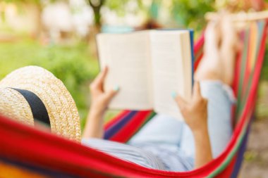 Yeşil bahçede rahat bir hamakta uzanmış kitap okuyan genç bir kadın..
