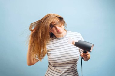 Güzel kadın güzel, uzun, ipeksi saçlarını kurutma makinesiyle mavi arka planda kurutuyor.