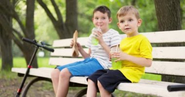 Çocuklar soda içer ve parkta yürüdükten sonra bankta oturarak sosisli yer. Yüksek kalite 4k görüntü