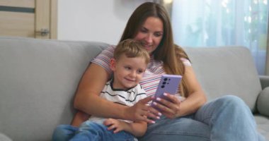 Kadın ve küçük oğlu akıllı telefonlu rahat koltukta oturuyorlar. Aileler evde birlikte vakit geçirirler. Akıllı telefon kullanır, oyun oynar, video izler, selfie çekerler. Yüksek kalite 4k görüntü