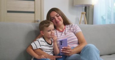 Kadın ve küçük oğlu akıllı telefonlu rahat koltukta oturuyorlar. Aileler evde birlikte vakit geçirirler. Akıllı telefon kullanır, oyun oynar, video izler, selfie çekerler. Yüksek kalite 4k görüntü