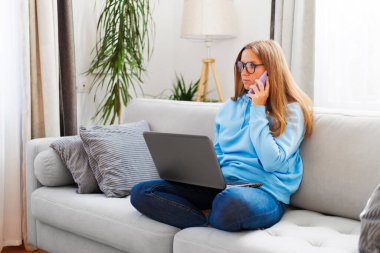 Ev ofisindeki kanepede otururken telefon ve laptop kullanan gözlüklü güzel bir kadın..