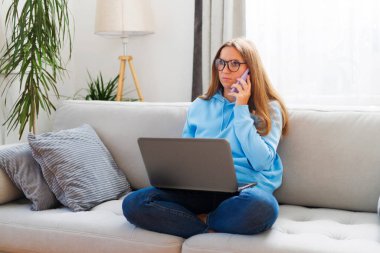 Ev ofisindeki kanepede otururken telefon ve laptop kullanan gözlüklü güzel bir kadın..