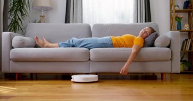 Robot elektrikli süpürge yerde çalışırken evdeki kanepede uyuyan çok yorgun bir kadın. Yüksek kalite 4k görüntü