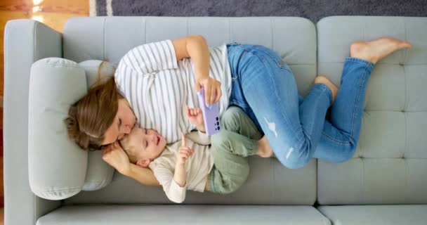 女人和她的小儿子躺在舒适的沙发上 带着智能手机 俯瞰四周 家人一起在家里用智能手机 玩游戏 看视频 高质量的4K镜头 — 图库视频影像