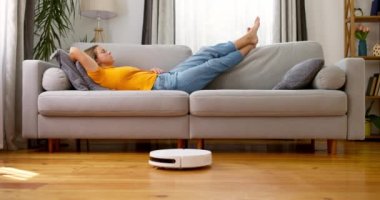 Robot elektrikli süpürge yerde çalışırken evdeki kanepede uyuyan çok yorgun bir kadın. Yüksek kalite 4k görüntü