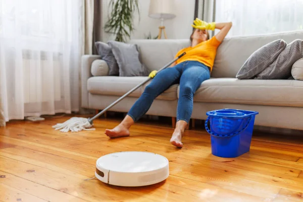Robotic vacuum cleaner cleaning a room while tired woman relaxing on the sofa after doing housework. Concept of the advantages of modern cleaning technologies, buying a robot vacuum cleaner