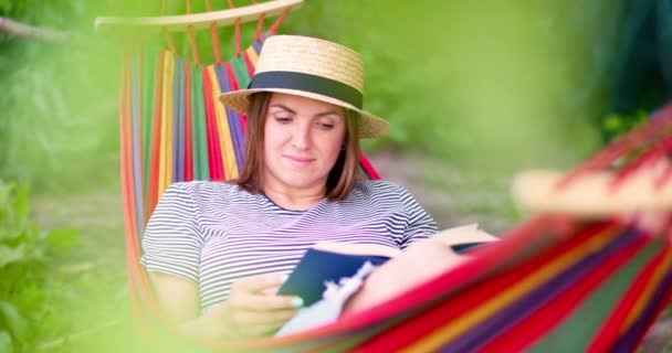 若いです女性読書本に横たわっている間に快適なハンモックで緑の庭 — ストック動画