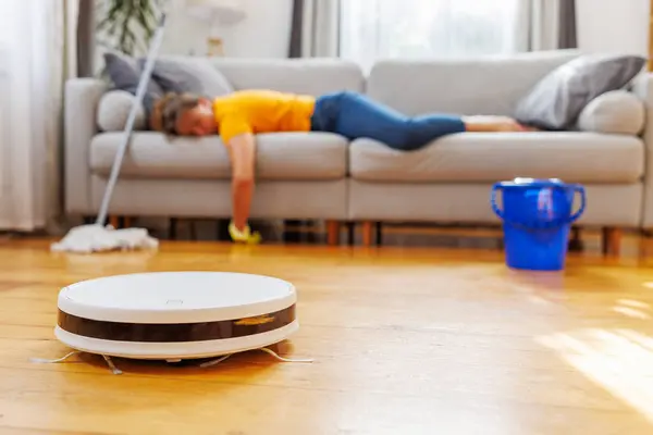Robotic vacuum cleaner cleaning a room while tired woman relaxing on the sofa after doing housework. Concept of the advantages of modern cleaning technologies, buying a robot vacuum cleaner