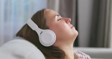 Genç bir kadın rahat bir kanepede, kulaklık takmış, kulaklık takmış, evde müzik ve müzik dinlemeyi seviyor. Yüksek kalite 4k görüntü