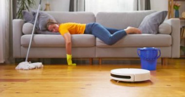 Sürekli temizlik yapmaktan bıkmış bir kadın rahat bir kanepede yatarken bir robot elektrikli süpürge temizlik yapıyor. Temizlik için yenilikçi teknolojiler. Yüksek kalite 4k görüntü