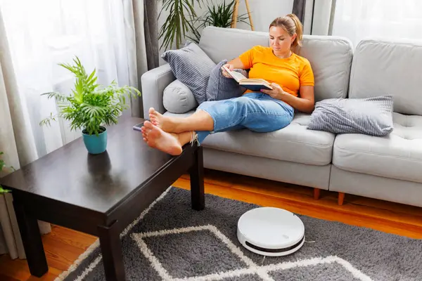 Robotic vacuum cleaner cleaning a room while a woman relaxing, reading book on the sofa. Innovative technologies for cleaning