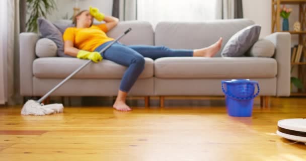 厌倦了不断的清洁 一个女人躺在舒适的沙发上 而一个机器人吸尘器做清洁 清洁的创新技术 高质量的4K镜头 — 图库视频影像