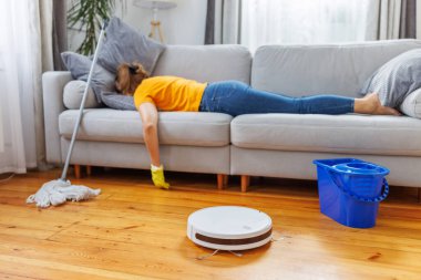 Ev işi yaptıktan sonra yorgun bir kadın kanepede dinlenirken robotik süpürge odayı temizliyor. Modern temizlik teknolojilerinin avantajları, bir robot elektrikli süpürge satın almak