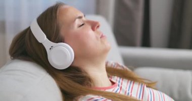 Genç bir kadın rahat bir kanepede, kulaklık takmış, kulaklık takmış, evde müzik ve müzik dinlemeyi seviyor. Yüksek kalite 4k görüntü