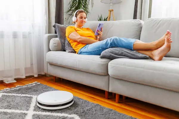 A woman relaxes on a comfortable sofa, immersed in music through headphones and a smartphone, while a robot vacuum cleans the room effortlessly