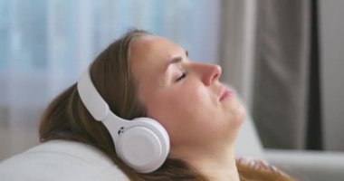 Genç bir kadın rahat bir kanepede, kulaklık takmış, kulaklık takmış, evde müzik ve müzik dinlemeyi seviyor. Yüksek kalite 4k görüntü
