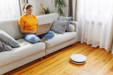 Kadın dizüstü bilgisayar kullanarak kanepede dinlenirken, robot süpürgesi odayı temizliyor..