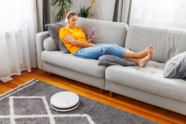 A woman relaxes on a comfortable sofa, immersed in music through headphones and a smartphone, while a robot vacuum cleans the room effortlessly
