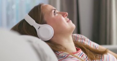Genç bir kadın rahat bir kanepede, kulaklık takmış, kulaklık takmış, evde müzik ve müzik dinlemeyi seviyor. Yüksek kalite 4k görüntü