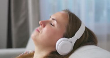 Genç bir kadın rahat bir kanepede, kulaklık takmış, kulaklık takmış, evde müzik ve müzik dinlemeyi seviyor. Yüksek kalite 4k görüntü