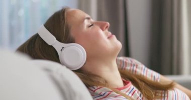 Genç bir kadın rahat bir kanepede, kulaklık takmış, kulaklık takmış, evde müzik ve müzik dinlemeyi seviyor. Yüksek kalite 4k görüntü