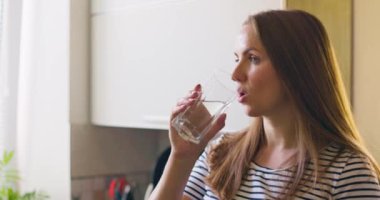 Bardak tutan, temiz, filtrelenmiş su içen yetişkin bir kadın ev mutfağında tek başına susuzluğunu gideriyor. Yüksek kalite 4k görüntü