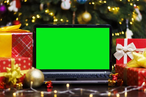 Laptop computer with green blank empty screen on table with gifts and garland on Christmas tree background. Mockup monitor