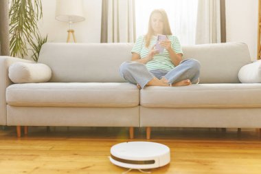 Kulaklık takan bir kadın rahat bir koltukta oturup, akıllı telefonundan müzik dinlerken bir robot süpürge temizlik yapıyor.