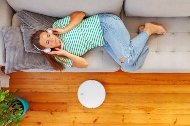 Kulaklık takan bir kadın rahat bir koltukta uzanıyor, dinleniyor ve müzik dinliyor. Robot elektrikli süpürge ise temizlik yapıyor.