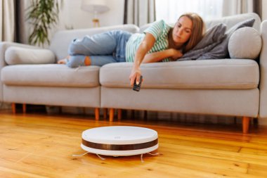 Bir kadın kanepede dinlenirken robot elektrikli süpürge odayı temizliyor. Bir kadın vakumu uzaktan kumandayla çalıştırıyor.