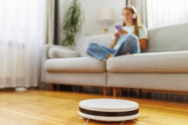 Kulaklık takan bir kadın rahat bir koltukta oturup, akıllı telefonundan müzik dinlerken bir robot süpürge temizlik yapıyor.