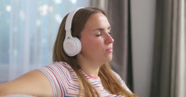 Kablosuz kulaklık takan beyaz bir kadın evdeki parlak bir odada kanepeye oturur, en sevdiği müziği dinler ve kanepede dans eder, kollarını hareket ettirir. Yüksek kalite 4k görüntü