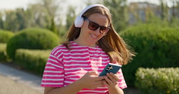 Χαμογελώντας Ανέμελη Γυναίκα Φορώντας Γυαλιά Ηλίου Και Λευκά Ασύρματα Ακουστικά — Αρχείο Βίντεο