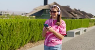 Güneş gözlüğü takan, kablosuz kulaklık takan, kaygısız gülümseyen bir kadın şehir parkında yürürken akıllı telefonunu kullanır. Yüksek kalite 4k görüntü