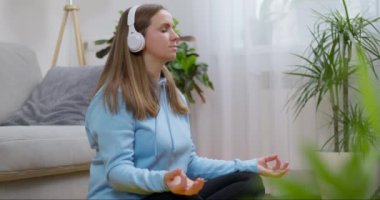 Kulaklık takıp evde yoga ve meditasyon yapan, oturma odasında poz verip gözleri kapalı rahatlamış mutlu bir genç kadın. Düşünceli meditasyon ve refah kavramı