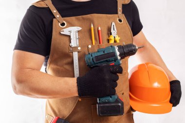 Önlüklü bir zanaatkar, elinde matkap ve miğfer tutuyor.