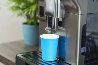 Mavi bir fincan, aydınlık ve çağdaş bir mutfakta taze bir sabah içkisi için hazırlanmış sofistike bir kahve makinesinin altında bekliyor..