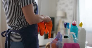  Çizgili gömlekli ve önlüklü biri turuncu temizleme eldivenleri giyiyor ve arka planda malzemeler var.
