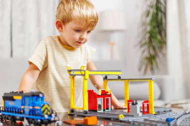 Odaklanmış genç bir çocuk renkli bir oyuncak tren seti ve kapalı alanda inşaat vinci ile oyun zamanının tadını çıkarıyor..