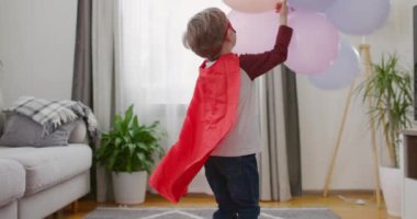 Süper kahraman kostümlü küçük bir çocuk rahat bir oturma odasında renkli balonlarla oynuyor. Yüksek kalite 4k görüntü