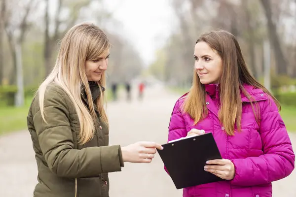 Two women engaged in a conversation with a clipboard on a park path.