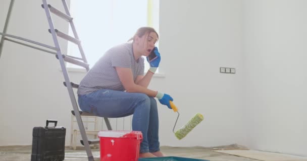 疲惫的女人坐在梯子上 手里拿着油漆滚筒在电话里交谈 可能需要专业的整修帮助 高质量的4K镜头 — 图库视频影像