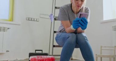 Merdivende oturan yorgun bir kadın, elinde boya silindiriyle telefonda konuşuyor, muhtemelen profesyonel yenileme yardımı istiyor. Yüksek kalite 4k görüntü