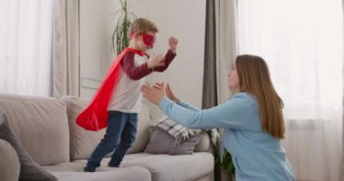 Süper kahraman kostümlü bir çocuk, parlak oturma odasında hevesli bir anneyle oynuyor. Yüksek kalite 4k görüntü