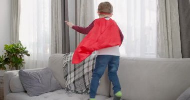 Evdeki kanepede zıplayan süper kahramanı oynayan genç çocuk. Kapalı mekan eğlencesi. Hayal gücü ve çocukluk kavramı. Yüksek kalite 4k görüntü